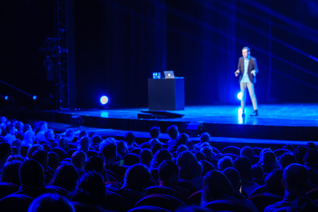 Yleisö seuraa puheenvuoroa hämärässä salissa, jossa sinisellä valaistulla lavalla esiintyjä seisoo pöydän ja kannettavan tietokoneen vieressä.