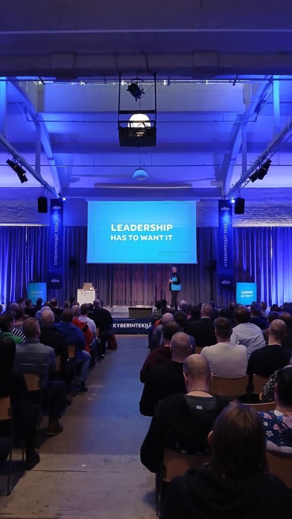 Yleisö seuraa kyberturvatapahtuman puheenvuoroa salissa, jossa sinisellä valaistulla lavalla esiintyjä seisoo suuren valkokankaan edessä. Valkokankaalla lukee “LEADERSHIP HAS TO WANT IT”, ja lavan sivuilla näkyvät Kyberintekijät 2026 -tapahtuman bannerit.