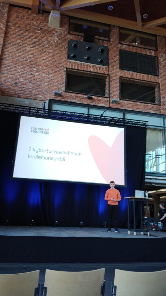 Puhuja seisoo lavalla tiilirakenteisessa tapahtumatilassa suuren esitysnäytön alla. Kankaalla näkyy otsikko “7 kyberturvaviestinnän kuolemansyntiä”, ja lavalla on myös korkea pöytä tietokoneineen.