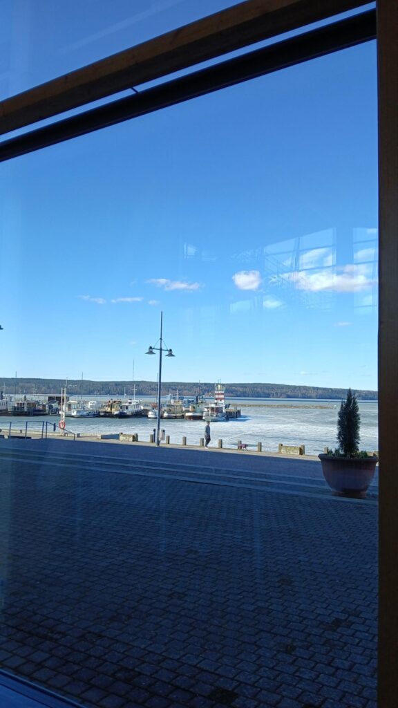 Näkymä suurten ikkunoiden läpi satama-alueelle, jossa laiturissa on useita aluksia ja osin jäässä oleva vesialue. Etualalla näkyy tapahtumatilan ikkunoiden heijastuksia sekä kivipäällysteinen piha.