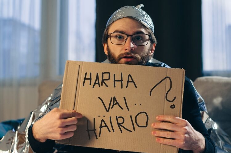 Harha vai häiriö webinaarisarja syksyllä 2026. Foliohattuinen mies istuu sohvalla ja pitelee kylttiä, jossa lukee Harha vai häiriö.