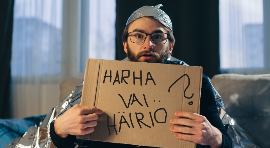Harha vai häiriö webinaarisarja syksyllä 2026. Foliohattuinen mies istuu sohvalla ja pitelee kylttiä, jossa lukee Harha vai häiriö.